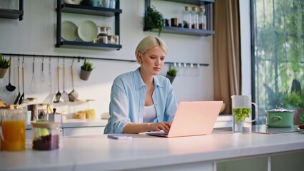 Focused blonde working laptop sitting kitchen table closeup. Woman sipping tea