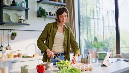 Joyful brunette listening music in earbuds stirring vegetables in kitchen area