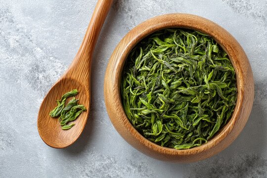 Wooden bowl filled with loose leaf green tea, beside a wooden spoon with tea leaves. - Powered by Adobe
