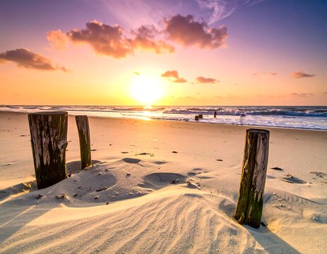 Sunrise over a tranquil beach, three weathered wooden posts stand in the sand, waves gently lapping the shore