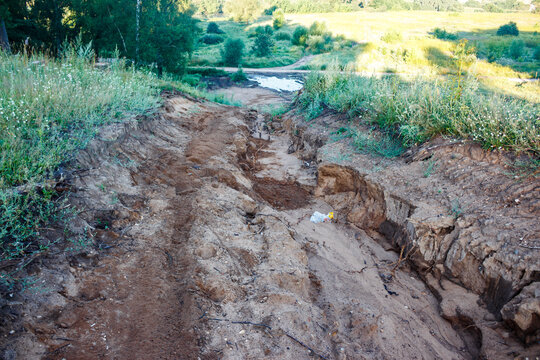 Deep rain-washed sandy gully, exhibiting severe erosion patterns through a rustic landscape. Wild grass borders steep banks leading to a small puddle below