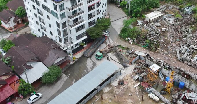 From a high angle, a team of laborers is handling a large pile of recently delivered steel beams within a construction yard that sits immediately adjacent to a modern apartment building