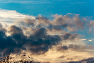 Dynamic evening sky with a vibrant array of clouds. Golden hues blend with deep blues and grays, creating a striking contrast. A contrail streaks across the vast expanse