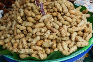 A close-up of a large mound of boiled peanuts in the shell, displayed in a blue basin lined with green leaves, with one opened pod showing purple nuts.