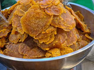 A close-up of a large, overflowing metal bowl filled with irregular, flattened pieces of dried candied mango (a traditional snack).
