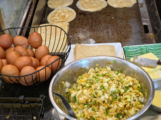 Overhead view of a cooking setup for Roti or Murtabak, with a bowl of egg and onion filling, a basket of eggs, and flatbreads cooking on a griddle.