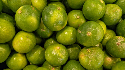 A close-up, overhead view of a large, tightly packed pile of whole, bright green limes with shiny skin, creating a vibrant, dense texture. 16:9 aspect ratio.