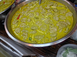 A large metal pot filled with Thai green curry (Gaeng Keow Wan) containing numerous fish balls and bamboo shoots in a rich, greenish-yellow coconut milk sauce
