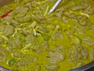 Close-up of a vibrant green curry (Gaeng Keow Wan) generously filled with fish balls, pieces of eggplant, and bamboo shoots, with a metal utensil visible in the upper right.