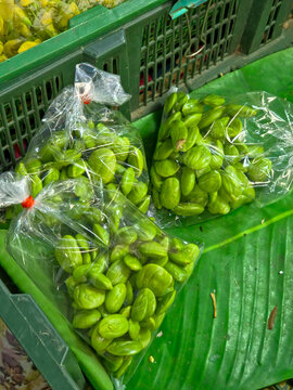 Three clear plastic bags filled with bright green, shelled bitter bean seeds (likely 'Sataw' or 'Parkia speciosa'), resting on a large green leaf near a green plastic crate.
