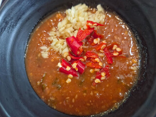 A top-down close-up of a dark bowl containing a thick, reddish-brown suki sauce dipping sauce generously topped with mounds of fresh minced garlic and bright red chili slices.
