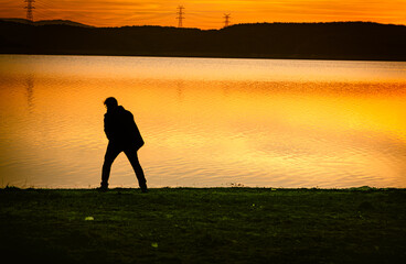 A single unrecognizable lonely man, wandering near a lake at sunset, yellow light shining on water