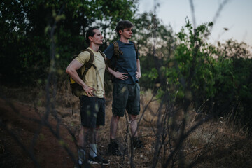 Naklejka premium Two men wearing hiking gear pause on a rugged forest path, ready for outdoor adventure, teamwork, and exploration amid trees and dry grass.