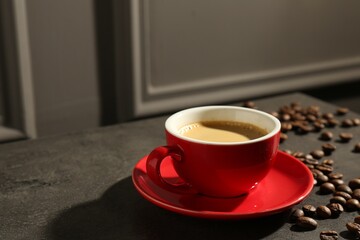 Red cup of aromatic coffee, saucer and beans on dark textured table, closeup. Space for text