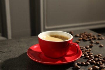 Red cup of aromatic coffee, saucer and beans on dark textured table, closeup