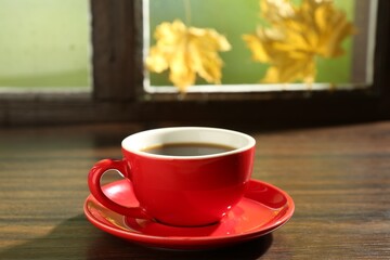Red cup of aromatic coffee and saucer on wooden sill near window, closeup