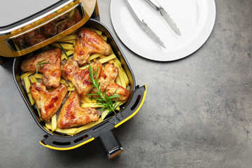 Air fryer with grilled chicken wings, french fries, rosemary and tongs on grey table, flat lay. Space for text