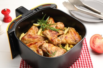 Grilled chicken wings with french fries in air fryer basket, tomato and tongs on white wooden table, closeup