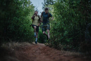 Obraz premium Two men with backpacks sprint along a dirt forest path, capturing energy and teamwork in an outdoor adventure. Ideal for stock listings about fitness, friendship, nature, and exploration.