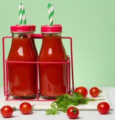 Glass bottles with red caps filled with tomato juice in a red metal stand. Fresh cherry tomatoes and a sprig of greenery lie nearby