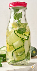 Glass bottles with red lids, filled with detox water containing cucumber and lemon slices and mint leaves, white table
