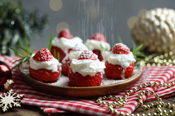 Sprinkling powdered sugar onto santa hats made of strawberries, whipped cream, rosemary and Christmas decor at wooden table against background with blurred lights, closeup. Bokeh effect