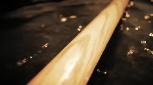 A close-up, angled view of a wooden baseball bat resting on a dark, reflective surface with scattered light reflections.