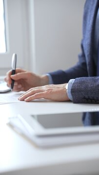 Close up of businessman's hands signing a contract with elegant pen, wearing a blue suit and a wristwatch, sitting at a white desk. Business people concept