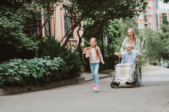 Heartwarming family moment as a girl runs beside grandpa and mom pushing his wheelchair along a sunny city park path