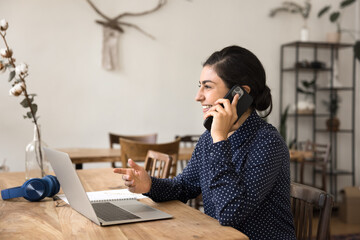 Smiling young businesswoman freelancer speak on smartphone before notebook