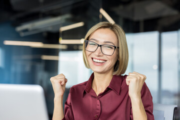 Happy businesswoman clenching fists in triumph, smiling broadly at a computer in a modern office,...
