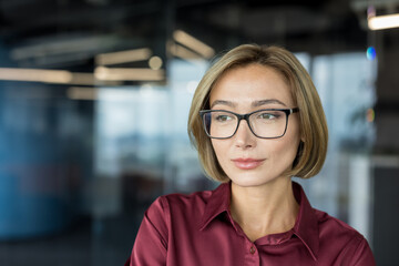 Professional businesswoman wearing glasses and a maroon shirt, confidently looking away in a modern...