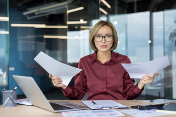 Businesswoman with glasses sitting at her office desk, holding two documents and looking directly...