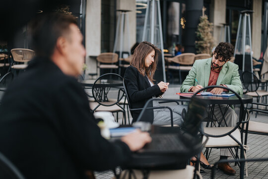 A casual outdoor cafe scene shows a man in a green blazer and a woman in black studying papers together, while a blurred coworker sips coffee in the foreground.