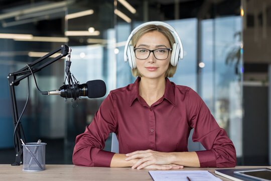 Professional woman wearing headphones and eyeglasses, sitting at a desk with a microphone, recording or streaming audio content in a modern office setup