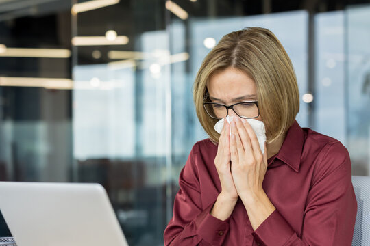 Young woman feeling unwell and blowing her nose with a tissue while working at her desk in a modern office, experiencing symptoms of cold, flu, or seasonal allergies