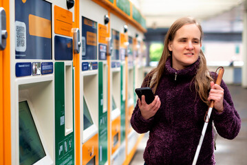 Visually impaired woman with white cane and smartphone standing at trains station and buying trains...