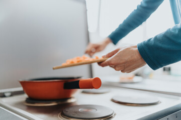 A view of a person preparing food in a domestic kitchen, adding ingredients to a cooking pan placed on a stove top, signifying home-cooked meals and cozy culinary practices.