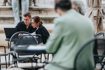 Two colleagues sit at an outdoor cafe table, focused on a laptop as they discuss documents. The scene conveys teamwork, planning, and casual collaboration in a relaxed setting.