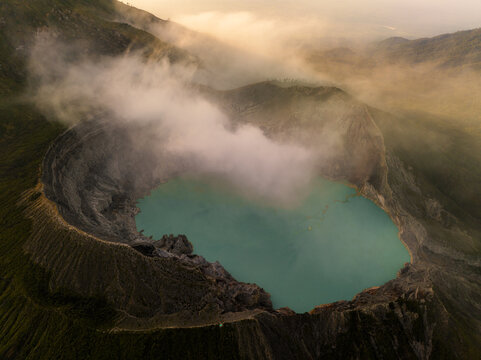 Aerial view of the stunning turquoise crater lake nestled within the volcanic caldera, shrouded in ethereal mist and rugged terrain, Mount Ijen, East Java, Indonesia.