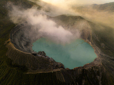 Aerial view of the breathtaking Ijen volcano crater lake, where turquoise waters meet rugged cliffs under a veil of ethereal mist, Mount Ijen, East Java, Indonesia.