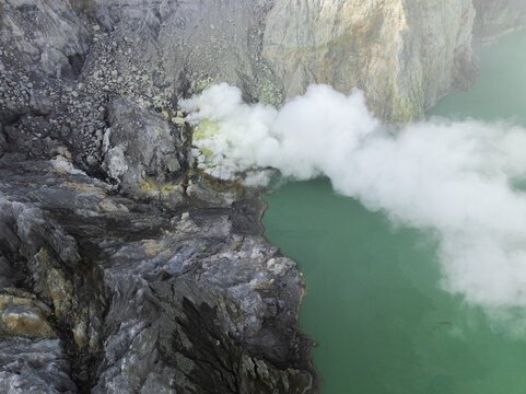 Aerial view of the Ijen volcano crater lake, its turquoise waters contrasting sharply with the rugged, sulfurous terrain and plumes of white smoke, Mount Ijen, East Java, Indonesia.