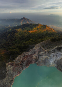 Aerial view of a turquoise lake nestled within a volcanic crater, rugged cliffs, and distant peaks bathed in golden light, Mount Ijen, East Java, Indonesia.