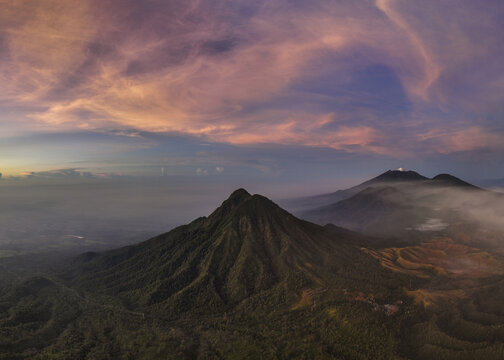 Aerial view of verdant peaks pierce through ethereal mists under a pastel sky, revealing the majestic Mount Ijen, East Java, Indonesia.