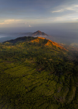 Aerial view of vibrant green forest contrasting with the warm, sunlit peak of Mount Ijen, East Java, Indonesia.