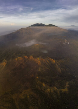 Aerial view of the majestic Mount Ijen, shrouded in mist and bathed in the warm glow of sunrise, casting long shadows across the rugged terrain, Mount Ijen, East Java, Indonesia.