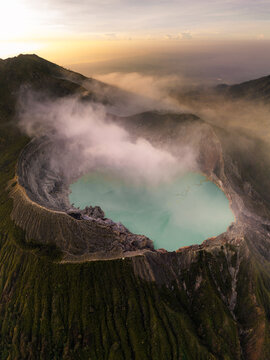 Aerial view of the Ijen volcano crater lake shrouded in ethereal mist, its turquoise waters contrasting with the rugged terrain, Mount Ijen, East Java, Indonesia.