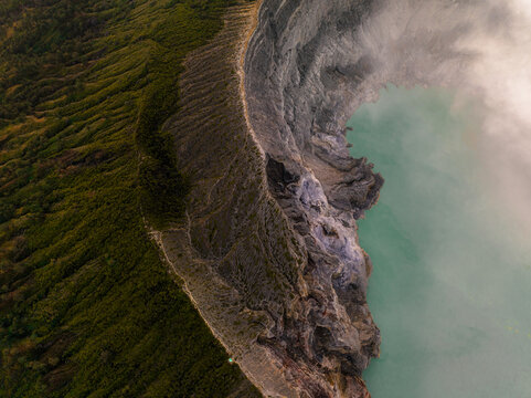 Aerial view of the rugged Ijen volcano crater lake, where turquoise waters meet the stark, rocky landscape under a veil of sulfuric mist, Mount Ijen, East Java, Indonesia.