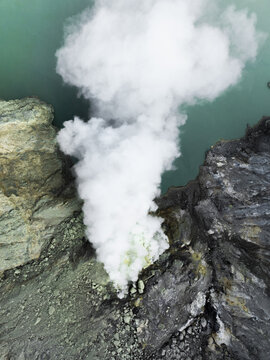 Aerial view of smoky plumes rising from the volcanic landscape, contrasting sharply with the turquoise crater lake, Mount Ijen, East Java, Indonesia.