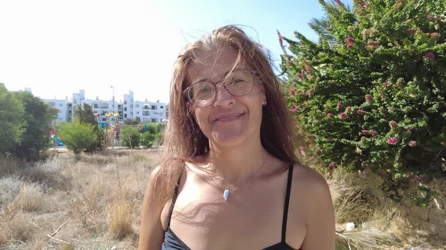 Middle-aged woman in glasses standing alone in the middle of the park looking at the camera. The female model in the video shot in the summer is wearing a strappy dress.